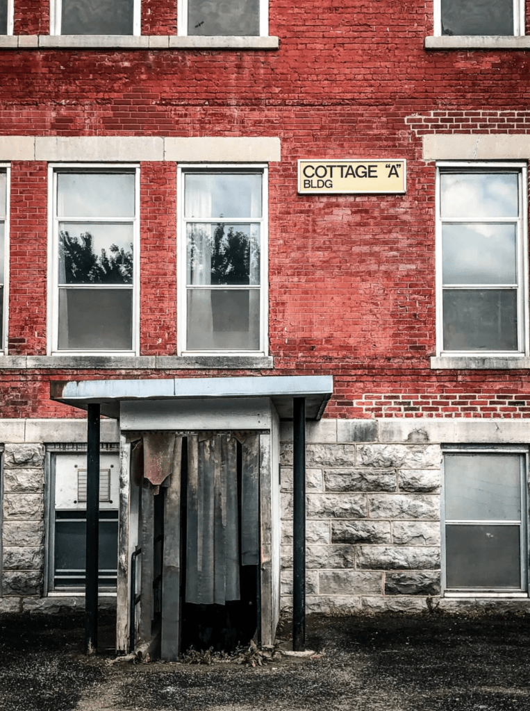 A colour photograph of the Cottage "A" building at the now-closed Huronia Regional Centre. The red bricks and concrete blocks of the building are worn away, and the plastic curtain on the entryway is torn. 