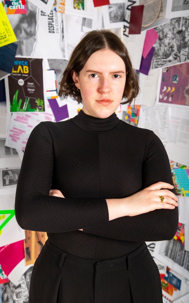 A portrait style photograph of a woman standing in front of a wall of colourful prints. She has short brown middle parted hair and is wearing a black turtleneck and trousers. She is standing with her arms crossed, looking at the camera.