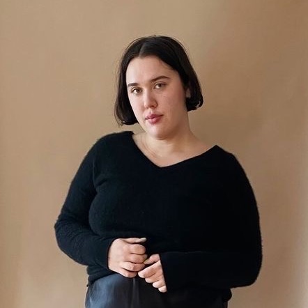 A portrait style photograph of a woman standing in front of a brown background. She is wearing a black cashmere sweater and a silky skirt. She is standing with her hands clasped and smiling at the camera with her head tilted to the side.