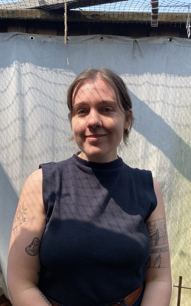 A portrait style photograph of a woman standing against a white backdrop. She has light brown hair pulled back into a bun and she is wearing a black high-necked tank top. She has visible black-and-white tattoos on her arms.
