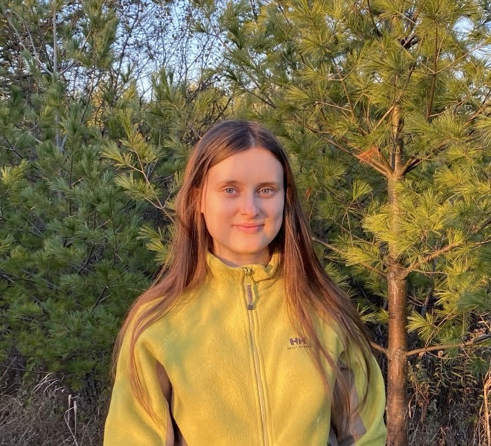 A portrait-style photograph of a person standing in front of several small pine trees. They have long, middle parted light brown hair, and are wearing a bright yellow zip-up fleece. They are lit by the sun and smiling into the camera.