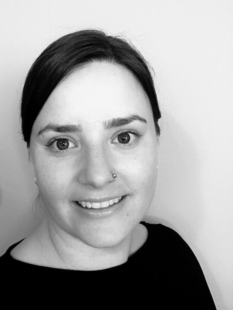 A black and white portrait style image of a woman smiling into the camera. She has dark hair pulled back into a bun, a silver nose ring, and a dark shirt. 
