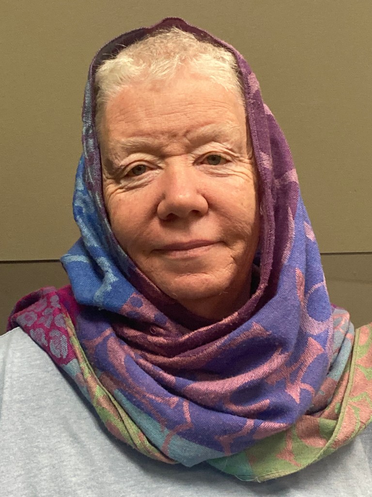 A portrait-style colour photograph of an older woman facing the camera. She is smiling gently. She has short, white hair, partly covered by a pashmina scarf wrapped around her head. The pashmina scarf is two-toned patchwork, in sections of pink on purple, green and peach, and dark blue and light blue. 