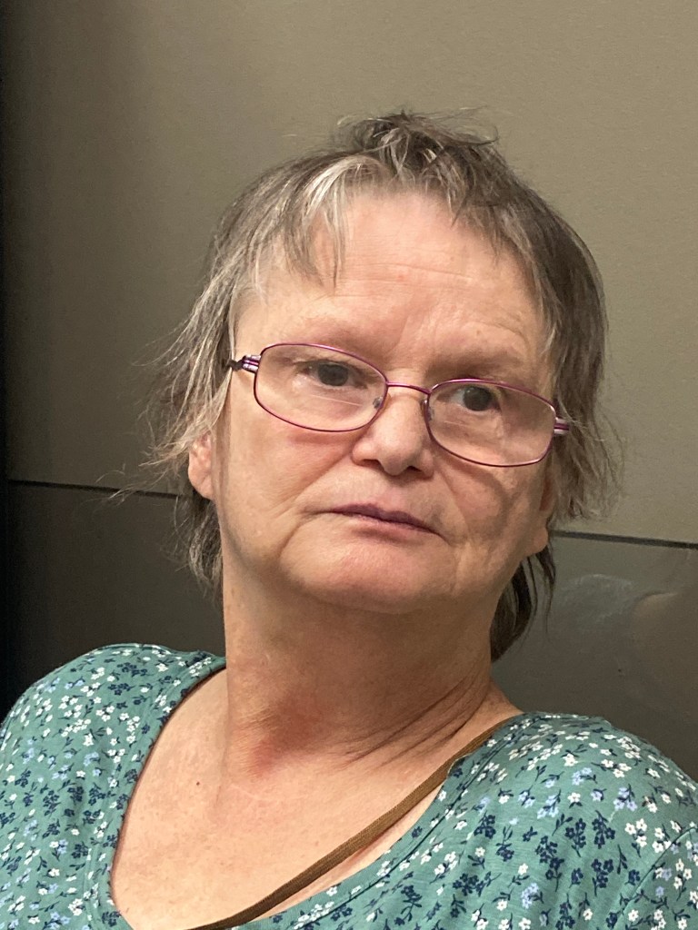 A portrait style colour photograph of an older woman looking off into the distance. She has chin-length grey hair, and is wearing thin wire-rimmed glasses, a scoop necked t-shirt covered in light blue flowers and a cross body bag. She has a solemn expression on her face. 
