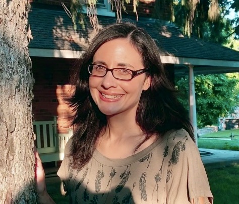 A portrait style photograph of a woman standing outside a red house. She has long, side parted brown hair, and is wearing thin, black glasses and a grey t-shirt with feathers printed on it. She is standing directly in the sunlight, smiling at the camera.