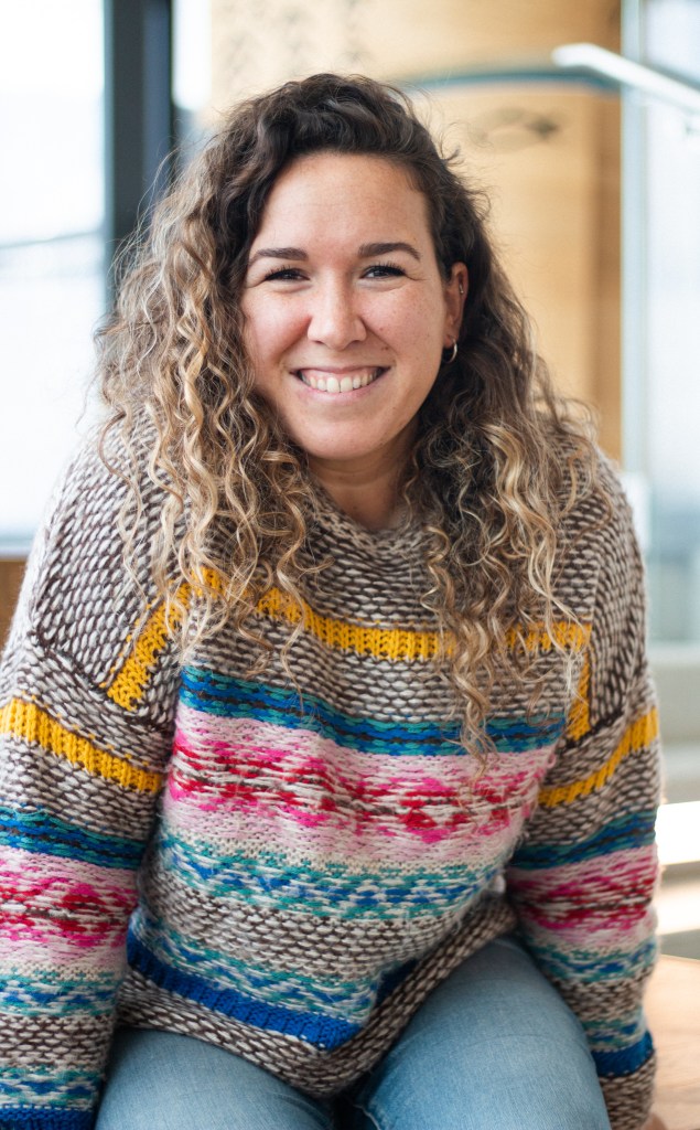 A portrait style photograph of a woman with curly, highlighted blonde hair. She is seated on a desk. She is wearing a hand-knit Fair Isle sweater in brown, yellow, blue and pinks, and a pair of jeans. She is smiling at the camera. 