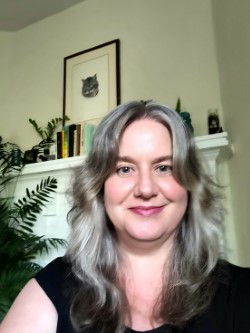 A selfie style portrait of a woman in front of a white fireplace and a background of foliage. She has shoulder length, middle parted silver hair and is wearing a black t-shirt. She is smiling at the camera. 